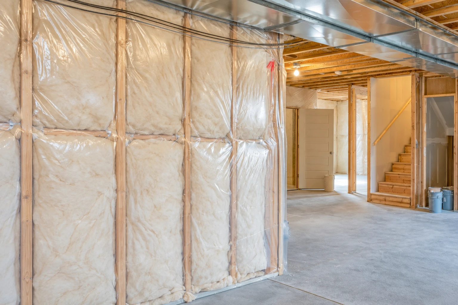 Basement with fiberglass insulation covered in clear plastic between wall studs, beside an unfinished concrete floor and wooden stairs.