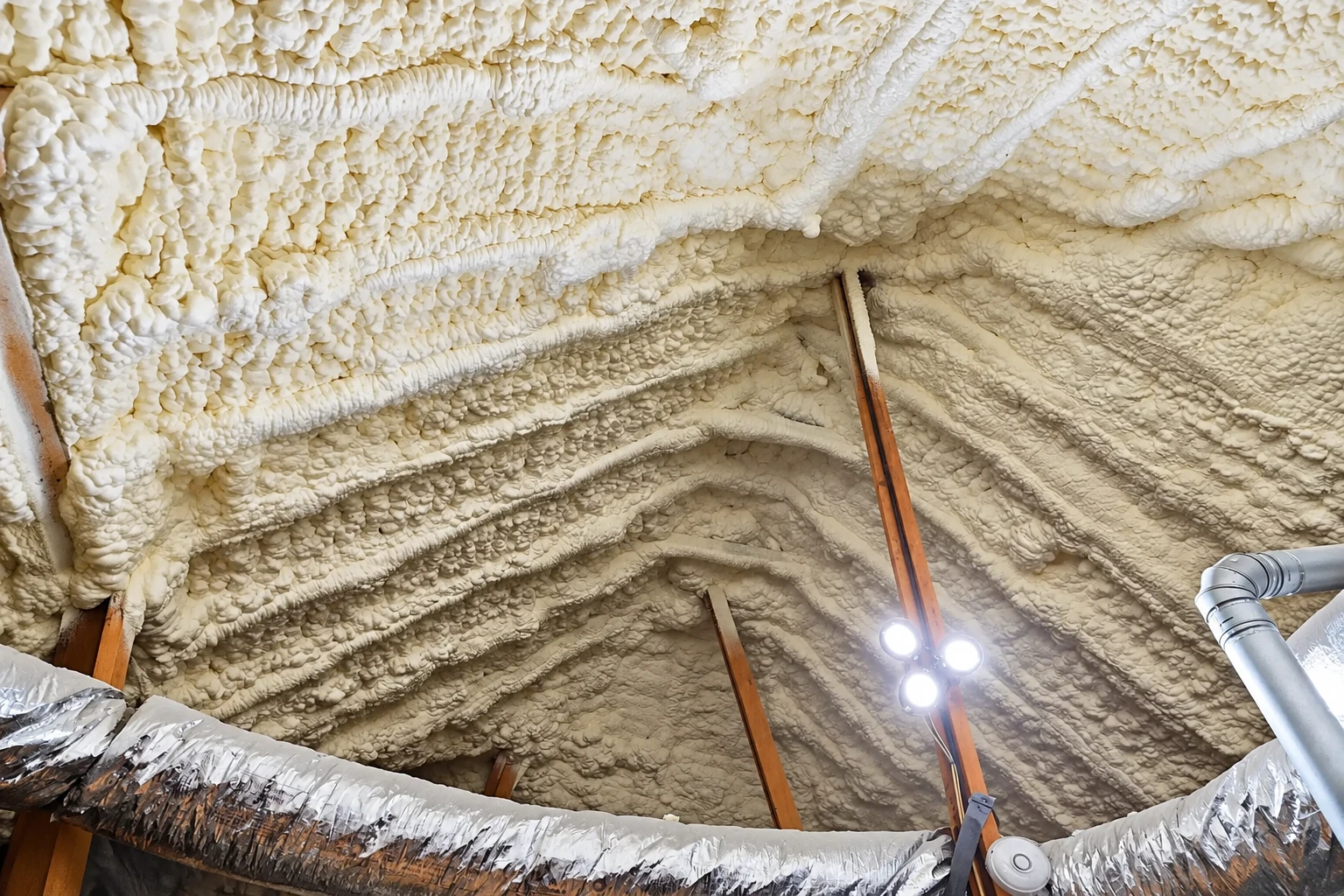 Close view of thick, textured white spray foam covering the roof and walls around exposed supports and ductwork.