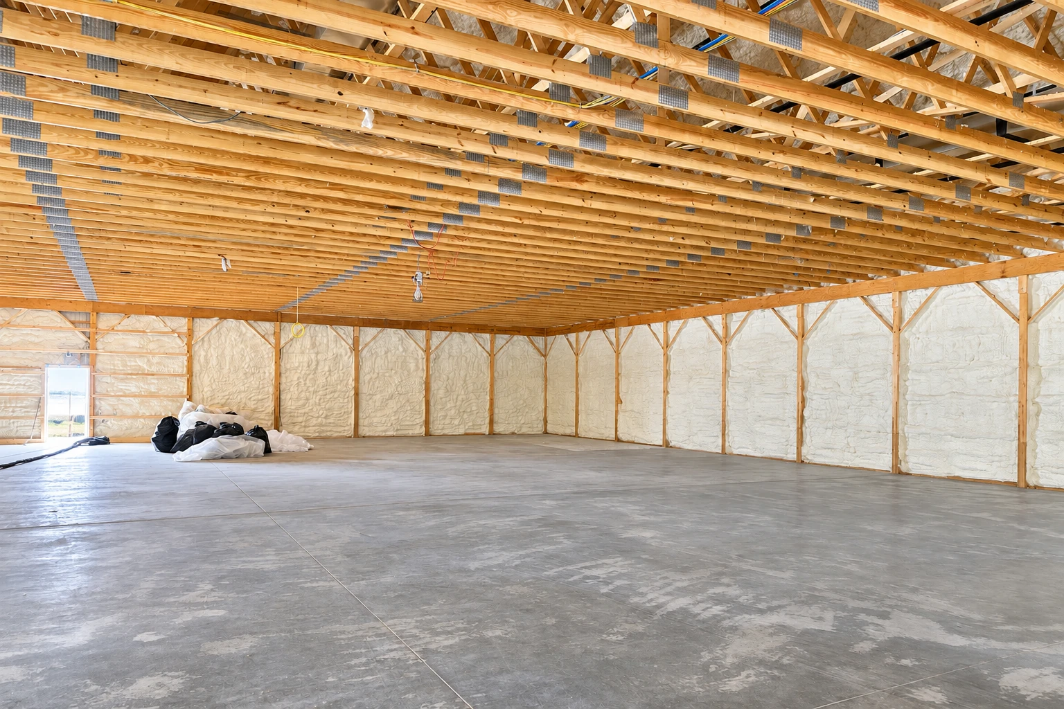 White spray foam lines the walls of a large commercial building with exposed wooden roof trusses and a concrete floor.