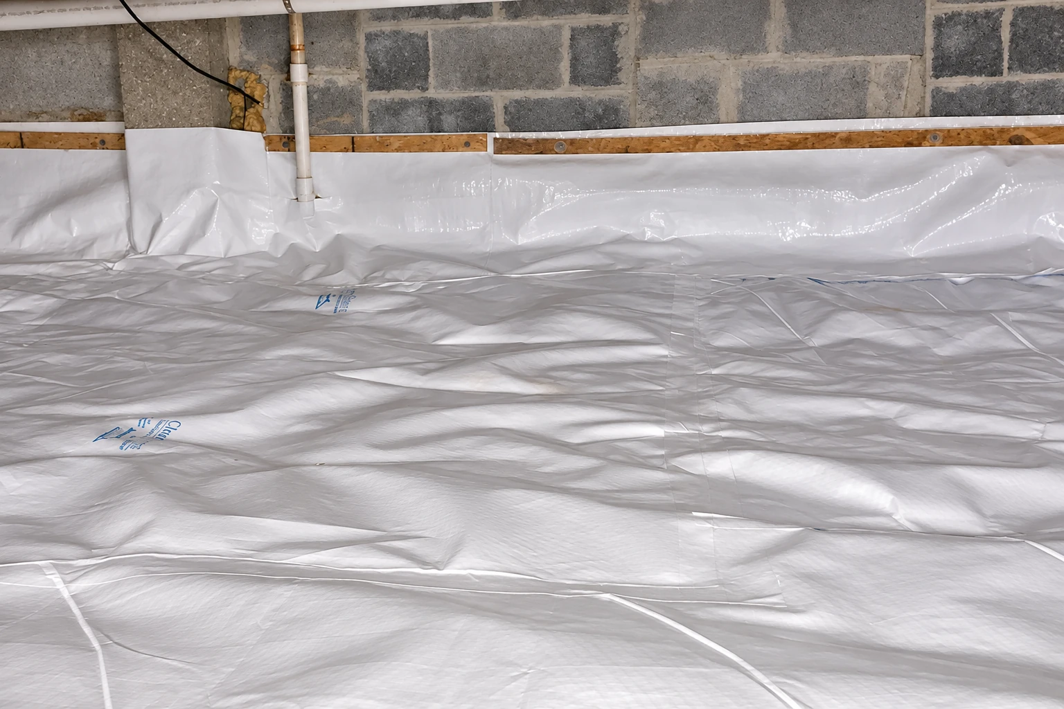 White vapor barrier sheeting covers the floor and lower walls of a crawl space with concrete block walls and plumbing.