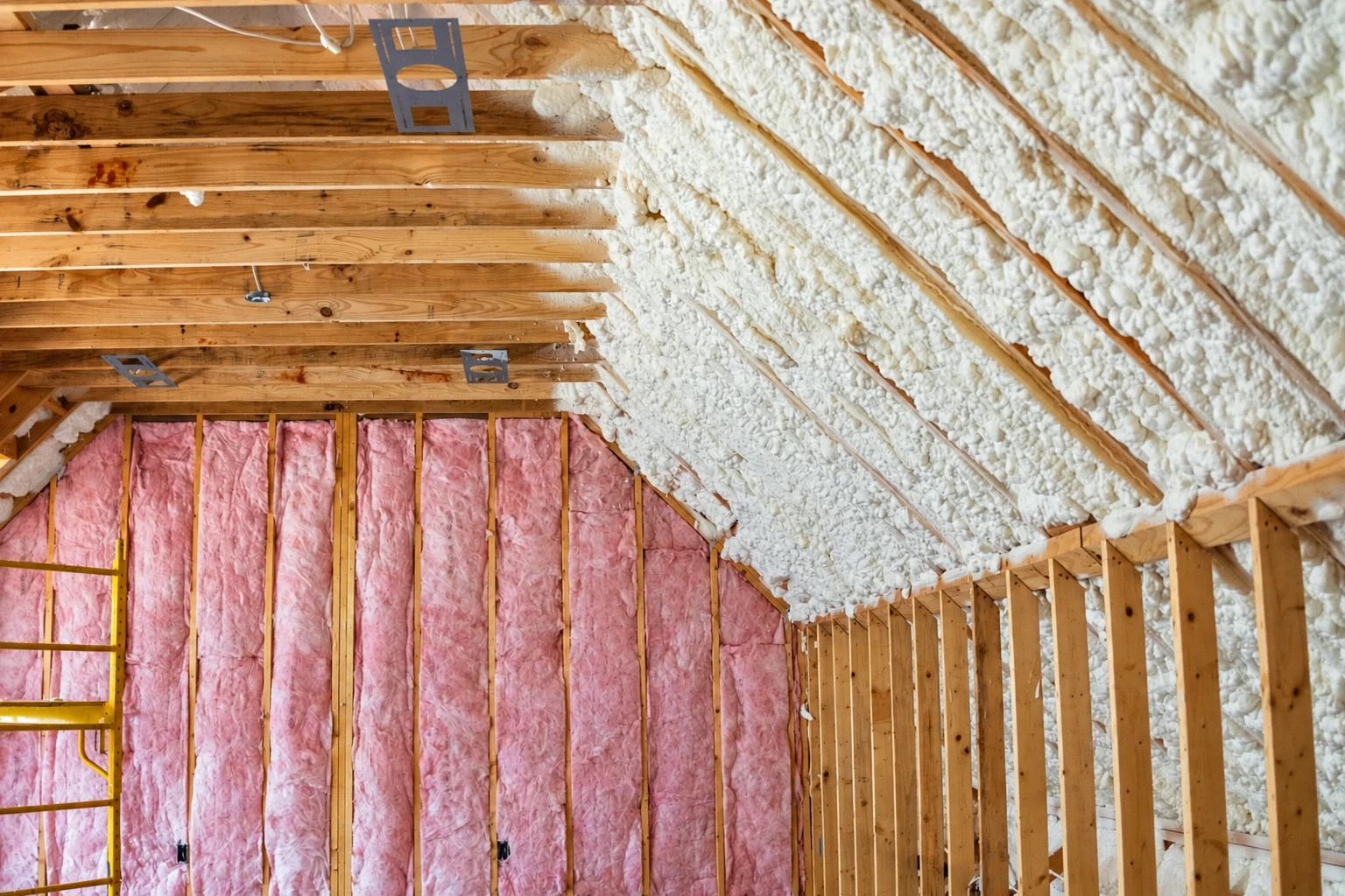 Open-cell spray foam coats the sloped roof framing above pink fiberglass batts in an unfinished attic.