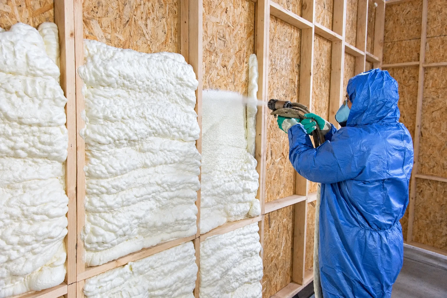 Worker in protective gear sprays white foam insulation into the open bays of a framed wall.