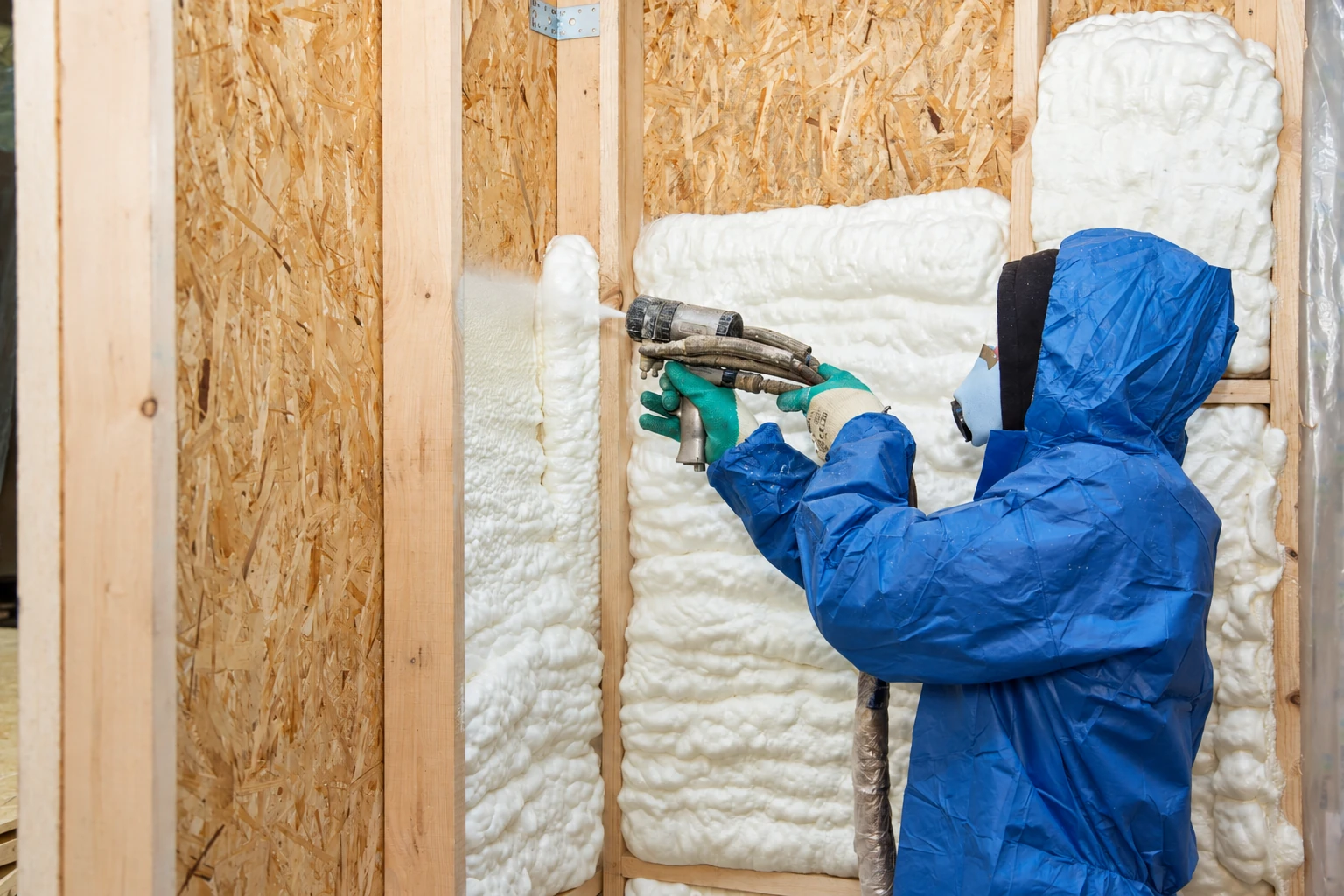 A worker in blue protective gear sprays foam insulation into the cavities of an unfinished framed wall.